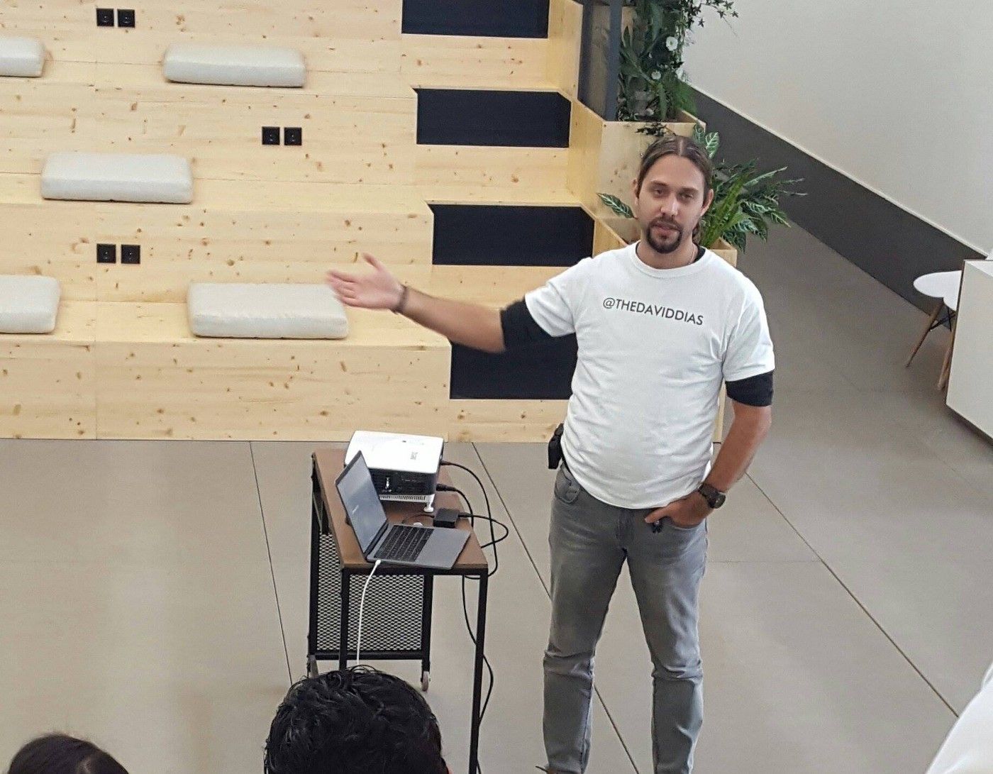 David Dias standing up and presenting near a video projector and MacBook. He is wearing a white tee-shirt with his username @thedaviddias printed in black.